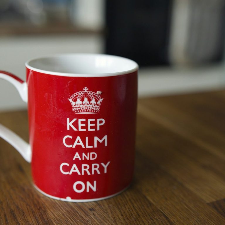 Rote Tasse mit dem Aufdruck "KEEP CALM AND CARRY ON" auf einem Holztisch.