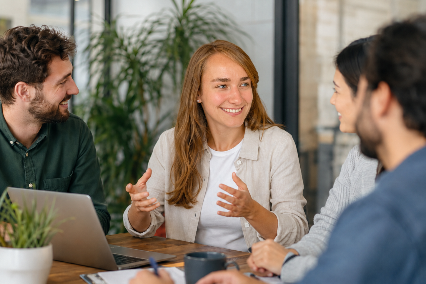 Business English Vier Personen in Geschäftsanzügen diskutieren an einem Tisch mit Laptops und Unterlagen.