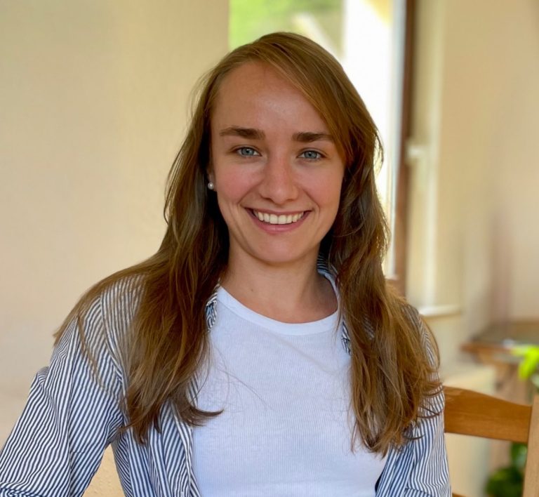 Junge Frau mit langen Haaren, sitzt lächelnd auf einem Stuhl in einem hellen Raum.