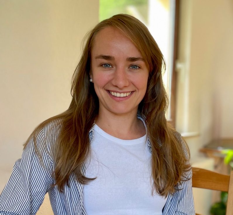 Junge Frau mit langen Haaren, sitzt lächelnd auf einem Stuhl in einem hellen Raum.